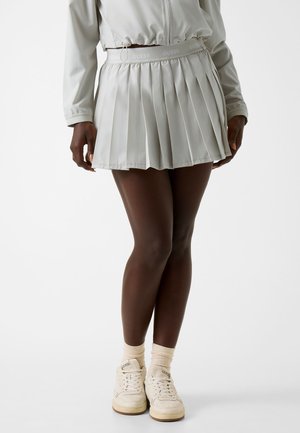 Modella che indossa una gonna plissettata grigio chiaro, giacca coordinata, calzini color crema e sneakers Guess crema, in piedi davanti a uno sfondo neutro.