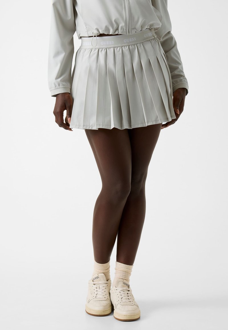 Mannequin portant une jupe plissée gris clair, une veste assortie, des chaussettes crème et des baskets Guess crème, debout sur un fond uni.