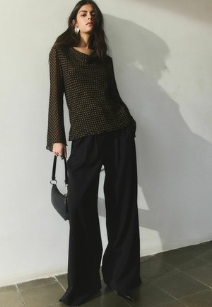 Kobieta w przezroczystej czarnej bluzce z długim rękawem ozdobionej żółtymi kropkami, szerokich czarnych spodniach, trzymająca czarną torebkę, stoi na tle jasnej ściany.