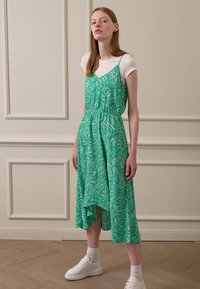 Vestido verde, com padrão, de corte solto, alças finas e bainha assimétrica, combinado com ténis brancos e uma t-shirt branca.