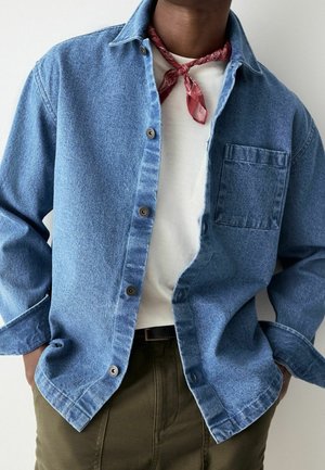 Personne portant une chemise en denim bleu déboutonnée sur un haut blanc, un bandana rouge noué autour du cou, et un pantalon vert olive les mains dans les poches.