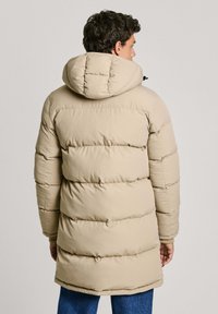 Beige puffer coat with a hood, featuring a quilted design and ribbed cuffs. Tailored fit extending to mid-thigh with a smooth texture.