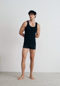 Un débardeur noir et un boxer ajusté sur un modèle masculin debout. Design simple, texture lisse, avec un fond uni et les pieds nus.