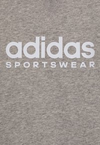 Šedé bavlněné tričko s výstřihem do V, velkým bílým logem "adidas" a textem "SPORTSWEAR", hladká textura a uvolněný styl.
