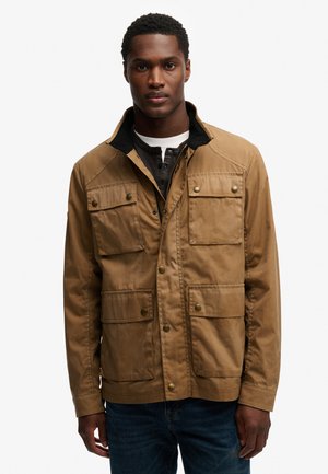 Mężczyzna noszący jasnobrązową kurtkę z czterema przednimi kieszeniami z patkami i mosiężnymi zatrzaskami, na ciemnej koszuli i dżinsach.