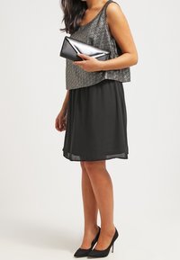 Černé bezramínkové šaty s vrstvenou, texturovanou stříbrnou vrchní částí; v kombinaci s černou kabelkou a černými vysokými podpatky. Hladká látka s volným střihem.