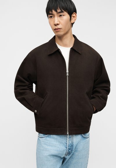 Νεαρός άνδρας που φοράει σκούρο καφέ μπουφάν με φερμουάρ και γιακά και ανοιχτόχρωμα μπλε τζιν, στέκεται με τα χέρια στις τσέπες του μπουφάν.