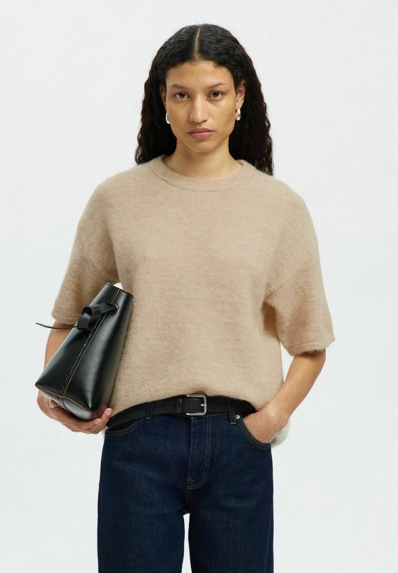 Pull en beige peluche à manches courtes, associé à un jean en denim bleu foncé. Sac à main noir angulaire avec un détail en nœud et des accessoires en argent.