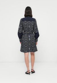 Woman standing with back to camera wearing a dark floral patterned long-sleeve dress with a belt and black heeled slingback shoes.