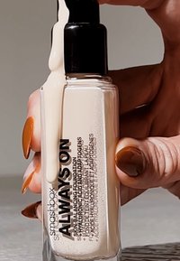 Flacon de fond de teint en verre transparent avec une pompe noire. Un liquide beige clair s'écoule du bec. Le texte de l'étiquette est en lettres noires sur le devant.