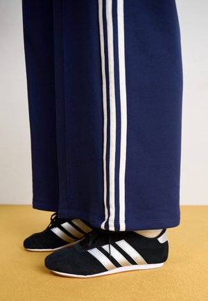 Pieds chaussés de baskets noires aux trois bandes blanches, associés à un pantalon bleu marine avec trois bandes blanches latérales, debout sur un sol moutarde.