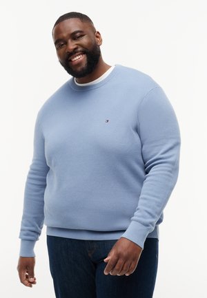 Uomo sorridente con capelli corti e barba che indossa un maglione azzurro chiaro texturizzato e jeans scuri su uno sfondo bianco uniforme.