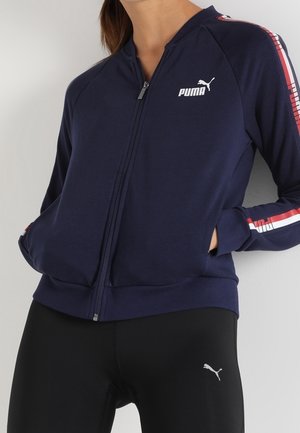 Osoba nosząca granatową kurtkę Puma zapinaną na zamek z czerwonymi i białymi paskami na rękawach oraz czarne legginsy Puma, ręce w kieszeniach kurtki.