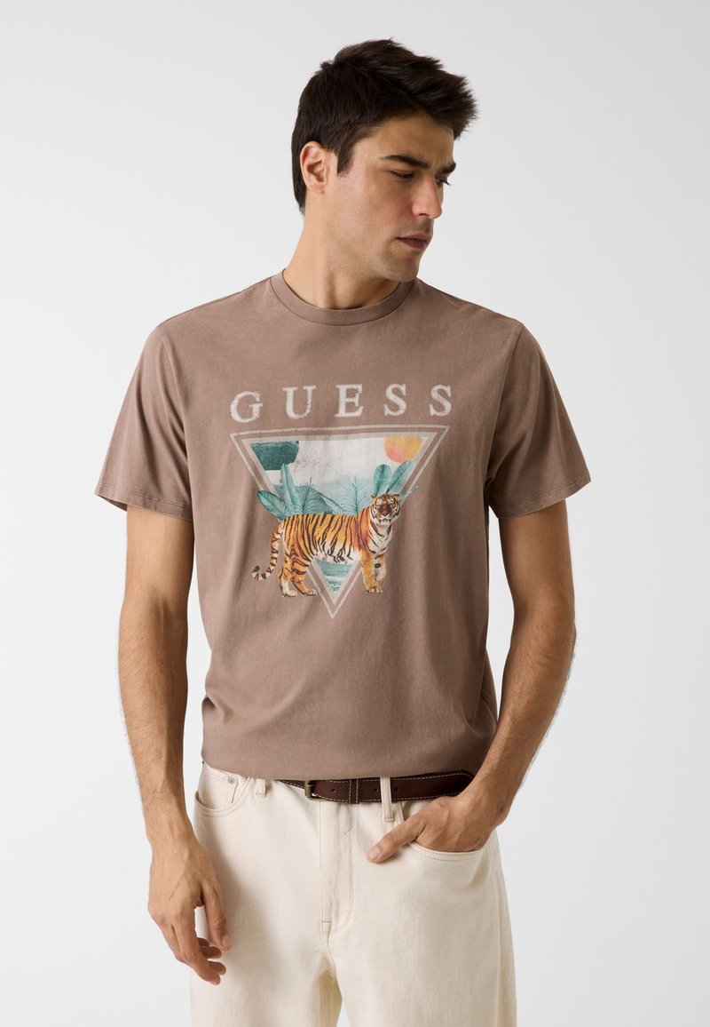 Άνδρας που φοράει καφέ μπλούζα Guess με γραφικό τίγρη και φύλλωμα, συνδυασμένη με λευκά παντελόνια και σκούρο ζωνάρι, κοιτάζει αριστερά.