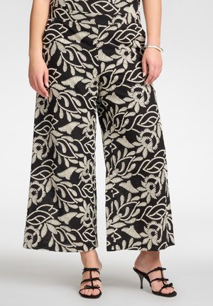 Zwarte broek met wijde pijpen en ingewikkelde witte blad- en wijnrankpatronen, gedragen met zwarte sandaalhakken met bandjes op een lichte achtergrond.