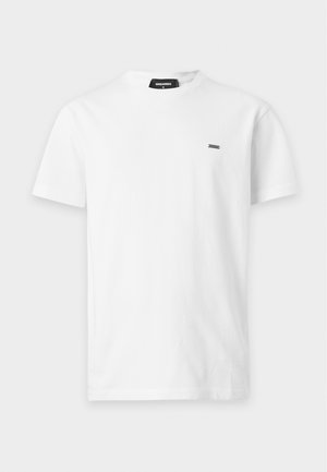 Uomo che indossa una t-shirt bianca a maniche corte, pantaloni grigi, scarpe nere e una collana a catena, in piedi con le mani in tasca.