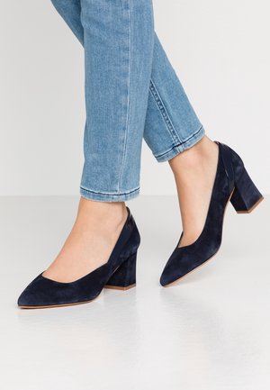 Marineblå ruskindspumpehøjhæle med spids tå og blokhæl, med en glat detalje ved hælen. Båret med lyseblå ankel-jeans.