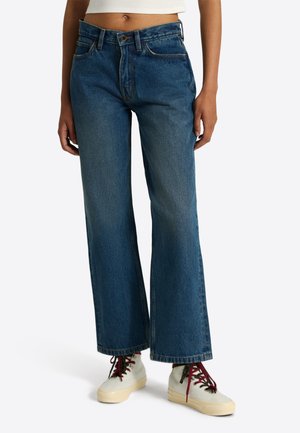 Femme portant un jean bleu taille haute à jambes larges avec bouton et fermeture éclair, associé à des baskets montantes blanches avec des lacets bordeaux.