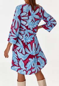 Kukkamekko, jossa on monivärinen kuvio sinisistä lehdistä viininpunaisella taustalla, kootussa vyötärössä ja kolmivartiset hihat.