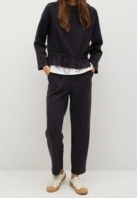 Mujer vestida con una camiseta negra de manga larga con cordones en la cintura, pantalones negros, zapatillas blancas y una camisa blanca debajo.