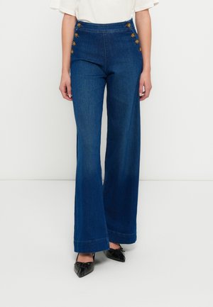 Vrouw draagt donkerblauwe, hooggetailleerde wijde jeans met zes messing knopen aan de voorkant, gecombineerd met zwarte puntige schoenen.