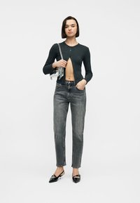 Femme portant un cardigan noir raccourci, un jean gris taille haute, des talons noirs pointus, tenant un sac à épaule transparent sur fond blanc.