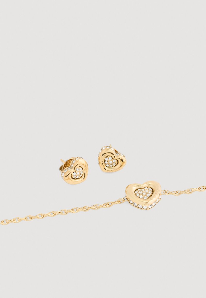 Set di gioielli dorati composto da orecchini a forma di cuore e bracciale, decorati con pietre trasparenti. Superficie liscia e dettagli di catena fine.