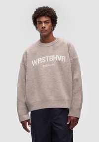 Beżowy, oversize sweter z miękkiego splotu, z białym logo "WRSTBHV" oraz "BERLIN" na klatce piersiowej, z prążkowanymi mankietami i dołem.
