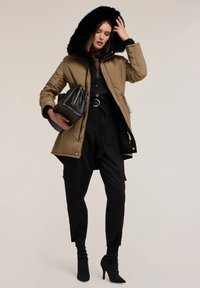 Giacca puffer tan con cappuccio in finto pelliccia nera, tasche con zip, abbinata a una camicia nera con botttoni e pantaloni, e una borsa nera.