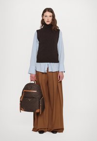 Un pulover brun fără mâneci, peste o cămașă cu dungi albastre deschise, pantaloni largi maro, ținând un rucsac cu un model în nuanțe de brun și bej.