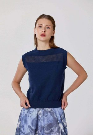 Femme aux cheveux châtain clair portant un top en maille bleu marine sans manches et une jupe à motif bleu-gris, debout, les mains sur les hanches, sur fond blanc.