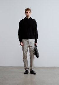 Sort ribstrikket sweater, lysegrå bukser og sorte sko. Model holder en sort taske. Minimalistisk design, glatte teksturer, neutrale farver.