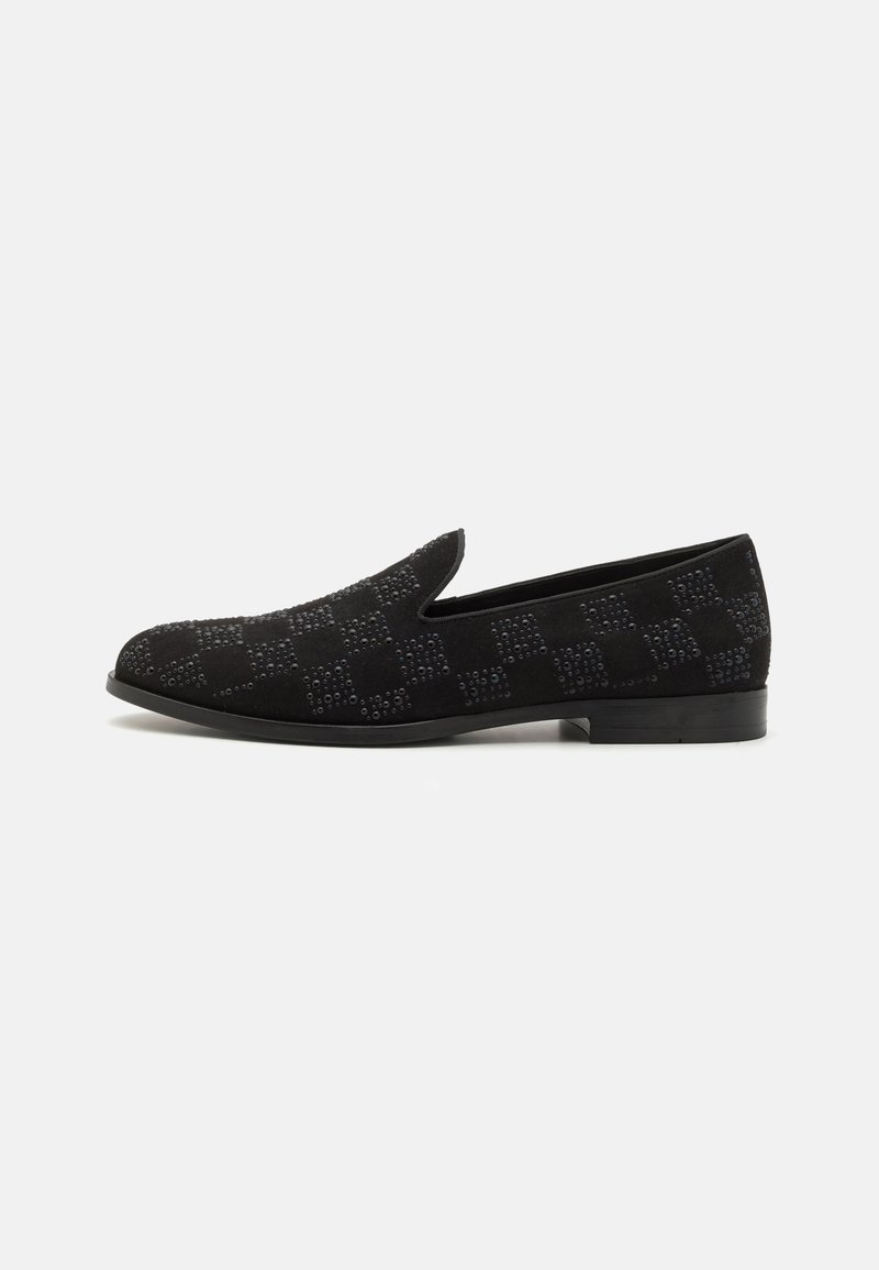 Mustmustest musta slip-on kinga, millel on tekstureeritud ruuduline muster väikeste studidega, madala musta kontsaga ja ümarate varvastega.