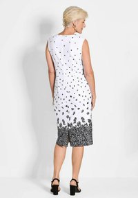 Ärmelloses weißes Kleid mit schwarzen floralen Mustern, die in einen intricaten schwarzen Saum übergehen. Glatte Textur, knielang, mit Reißverschluss hinten.