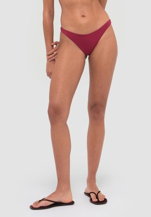 Kobieta w czerwonych majtkach od bikini i czarnych japonkach stoi na jednolitym jasnoszarym tle.