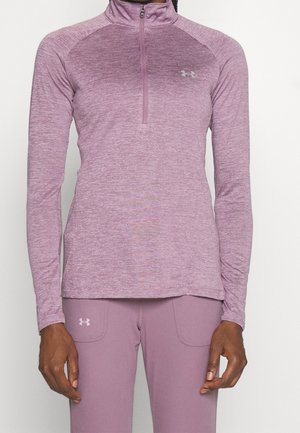 Frau trägt ein mauvefarbenes, langärmeliges Sportoberteil mit Viertelreißverschluss und passende Hose, Under Armour-Logos sind auf Brust und Oberschenkel sichtbar.