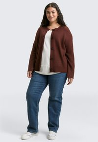 Bordeaux breimode cardigan met ronde hals, knopenluiting en lange mouwen, gecombineerd met een witte top en blauwe spijkerbroek, gedragen met witte sneakers.