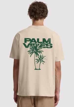 Osoba nosząca beżową koszulkę z zielonym napisem "PALM VIBES" i dwoma ilustracjami palm na plecach, stojąca na jednolitym tle.