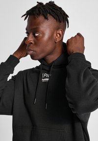 Svart hoodie med känguruficka, snörningar och en vit logotyp på bröstet. Har en mjuk textur och avslappnad passform.