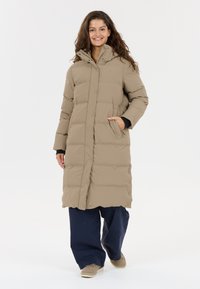 Beige pufferjakke med lang design, trykknapper og hætte. Den har sidelommer og bæres over navy bukser og lyse sko.