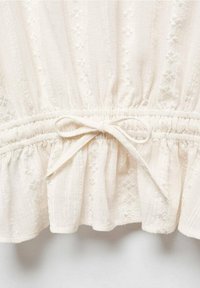 Blusa bianca a sbuffo con tessuto texturizzato, caratterizzata da un orlo arricciato e da un fiocco in vita. Leggeri motivi floreali sono visibili su tutta la superficie.