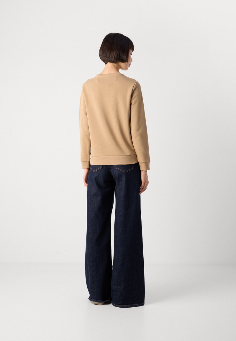 Person mit kurzen dunklen Haaren, die ein beiges Oberteil mit langen Ärmeln und eine dunkle weit geschnittene Jeans trägt, steht und schaut von hinten vor einem schlichten hellen Hintergrund.