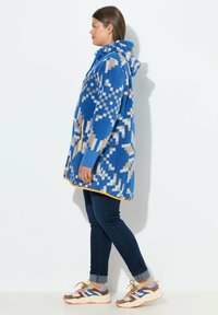 Felpa blu in pile con motivi geometrico in beige, rifiniture arancioni e maniche lunghe, abbinata a jeans scuri e sneakers multicolori.