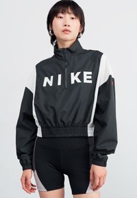 Coupe-vent noir Nike avec logo blanc, col zippé au milieu, taille élastique et panneaux latéraux blancs contrastants. Porté avec un short noir.