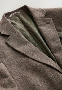 Braun strukturierter Blazer mit dezentem Karomuster, offen getragen, das ein grünes, gemustertes Futter zeigt. Auffällige Details sind eine einzelne grüne Akzentnaht.