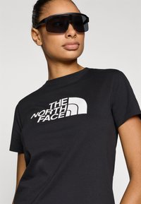 Μαύρο βαμβακερό μπλουζάκι με μεγάλο λευκό λογότυπο "The North Face" μπροστά. Το μοντέλο φοράει υπερμέγεθες μαύρα γυαλιά ηλίου.