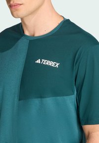 Camiseta deportiva de color teal con mangas cortas, que presenta un diseño en contraste, textura suave y un logo blanco de "TERREX" en el pecho.