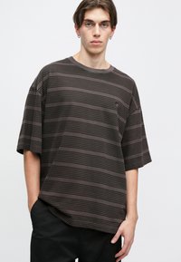 Camiseta de gran tamaño de color marrón oscuro con rayas, de mangas cortas y cuello redondo, que presenta una textura sutil y un pequeño detalle de logo en el pecho.