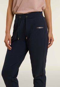 Pantaloni joggers blu navy con vita elasticizzata e lacci, caratterizzati da un logo metallico "FREDDY" sul lato sinistro e tasche laterali.