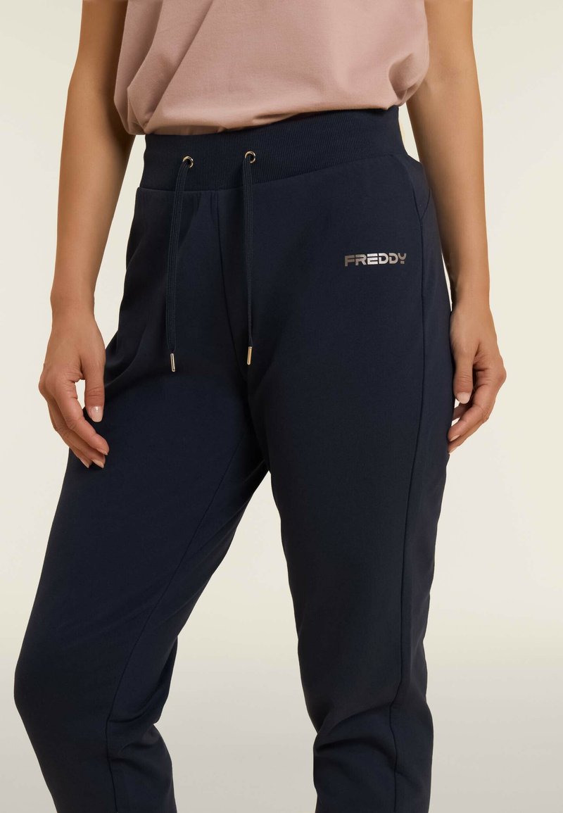 Pantaloni joggers blu navy con vita elasticizzata e lacci, caratterizzati da un logo metallico "FREDDY" sul lato sinistro e tasche laterali.